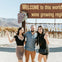 Three people posing and smiling in front of a sign that reads 'Welcome to this world famous wine growing region' with vineyards behind.