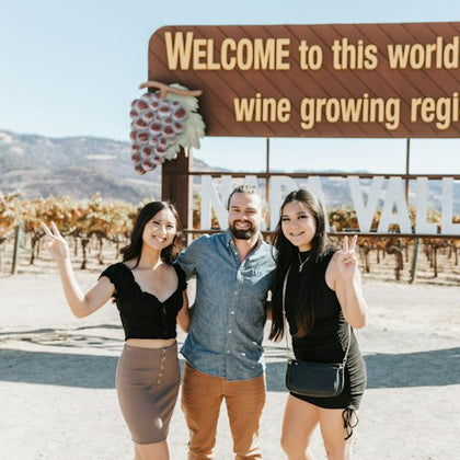 Three people posing and smiling in front of a sign that reads 'Welcome to this world famous wine growing region' with vineyards behind.