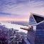 A modern skyscraper with a triangular observation deck, overlooking a cityscape and a river at sunset.