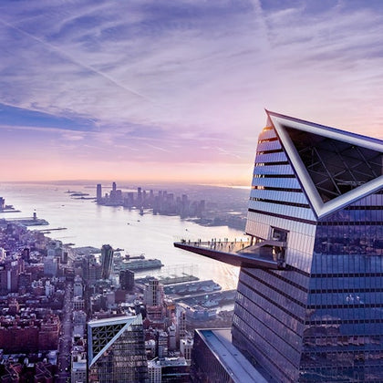 A modern skyscraper with a triangular observation deck, overlooking a cityscape and a river at sunset.
