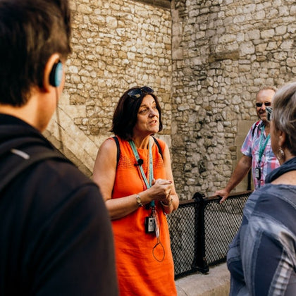A tour guide in an orange dress talks to a group of people in front of a stone wall.