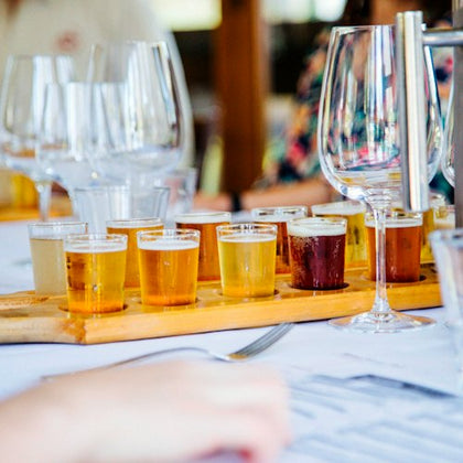 A flight of various beers in small glasses on a wooden tray, surrounded by wine glasses on a restaurant table.