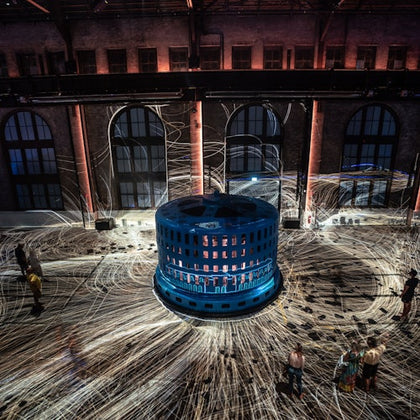 A large blue cylindrical installation emits light trails in a dimly lit industrial space with people observing.