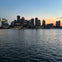 A city skyline at dusk reflected in calm water, with buildings lit against a transitioning orange and blue sky.