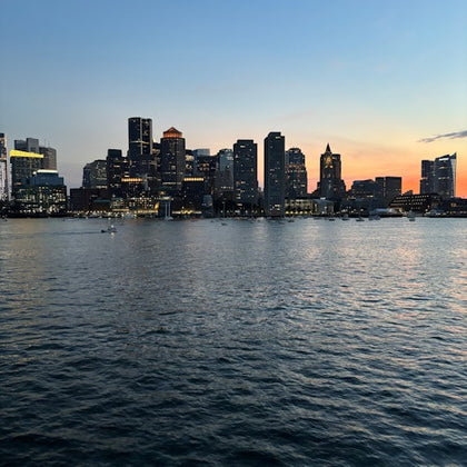 A city skyline at dusk reflected in calm water, with buildings lit against a transitioning orange and blue sky.