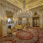 Ornate room with elaborate gold decorations, large chandelier, red patterned carpet, antique furniture, and decorative fireplace.