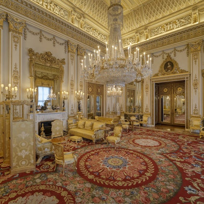 Ornate room with elaborate gold decorations, large chandelier, red patterned carpet, antique furniture, and decorative fireplace.