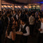 A crowded social gathering in an elegant indoor setting with large windows overlooking a cityscape at sunset.
