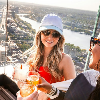 Two people toasting with cocktails on a rooftop with a cityscape and river in the background.