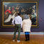 Two people stand in front of a large, ornate painting displayed in an art gallery, observing it attentively.