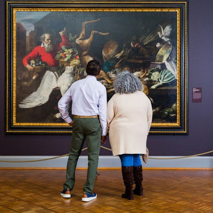 Two people stand in front of a large, ornate painting displayed in an art gallery, observing it attentively.