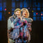 Two performers on stage, one embracing the other from behind, wearing vibrant costumes under colorful stage lighting.