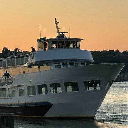 A white ferry boat on the water at sunset, with passengers visible on the deck. Background features a shoreline with trees.