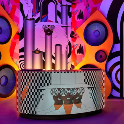 A colorful art installation featuring cylindrical structures, abstract patterns, and stylized human faces on a checkered counter.
