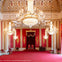 Opulent room with red walls, ornate chandeliers, golden accents, and two regal chairs on a platform under a canopy.