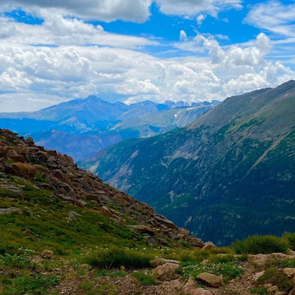 Mountain landscape with rocky terrain, green slopes, and distant peaks under a partly cloudy blue sky.