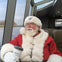 A man dressed as Santa Claus holding a branded coffee cup while seated inside a boat with a view of a river through the windows.