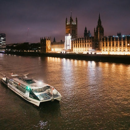 A lit river cruise boat travels on the Thames at night, passing by the illuminated Houses of Parliament.
