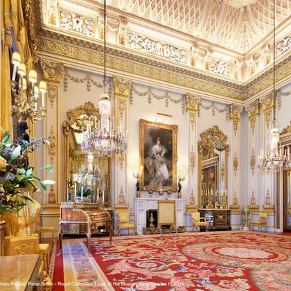 A lavish, ornate room with golden accents, chandeliers, a large portrait, and an intricate red and gold carpet.