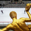 A gold statue overlooks an ice rink with skaters, surrounded by a stone wall and stairs with seated spectators.