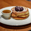 A stack of pancakes topped with berry compote and powdered sugar on a white plate, served with a small cup of syrup.