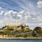 A historic building with a lighthouse sits atop a rocky hill surrounded by greenery, under a cloudy sky by the water.