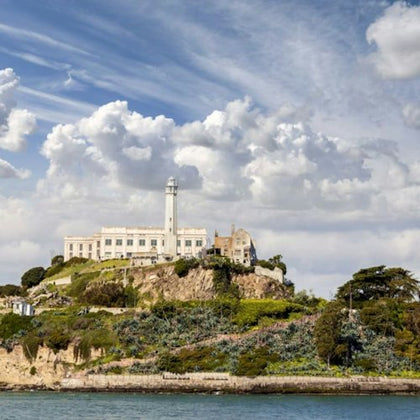A historic building with a lighthouse sits atop a rocky hill surrounded by greenery, under a cloudy sky by the water.