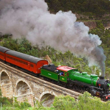 A green steam locomotive pulling red passenger cars crosses a stone bridge surrounded by lush greenery.