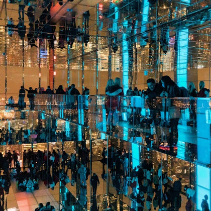 A mirrored room with people exploring, illuminated by blue and warm lights, creating endless reflections.