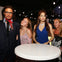 Three people pose with wax figures of a man in a suit and glasses and a woman in a blue dress, in a setting with string lights.