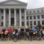 A group of people with bicycles standing in front of a large classical-style building with columns.