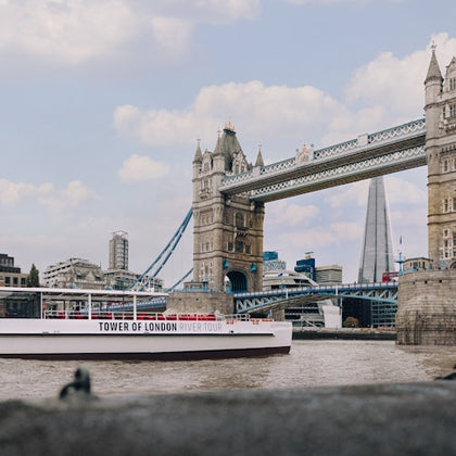 A view of Tower Bridge with a tour boat labeled 'Tower of London River Tour' on the Thames and the Shard in the background.