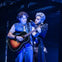 Two performers on stage, one holding a guitar, the other gesturing towards them, under blue lighting.