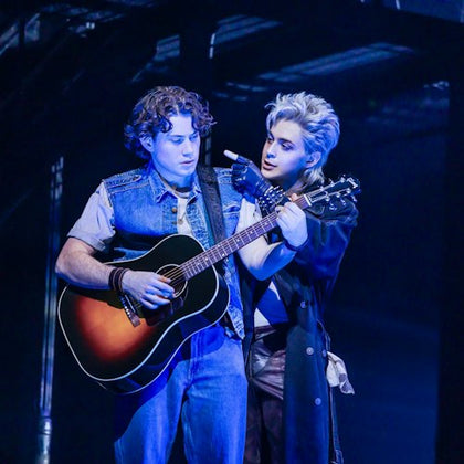 Two performers on stage, one holding a guitar, the other gesturing towards them, under blue lighting.
