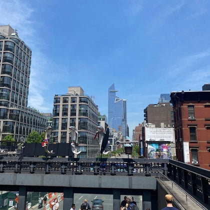 A view of modern buildings seen from an elevated walkway with sculptures and people enjoying the sunny day.