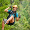 A person wearing a helmet and harness is ziplining through a forested area while smiling and gesturing with their hand.