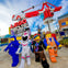Four LEGO character mascots stand under the LEGO Movie World entrance sign, with a themed amusement park in the background.