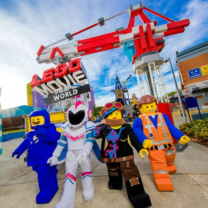 Four LEGO character mascots stand under the LEGO Movie World entrance sign, with a themed amusement park in the background.