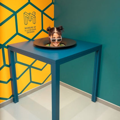 A person appears to have only their head visible on a blue table, surrounded by a yellow and teal geometric background.