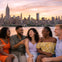 A group of friends laughing together on a waterfront with a city skyline and sunset in the background.
