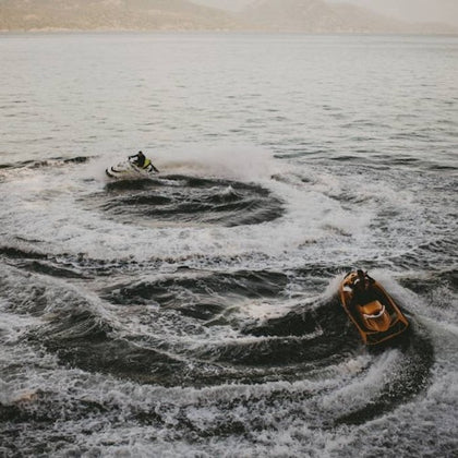 Two people riding jet skis, creating circular waves on a calm lake with mountains in the background.