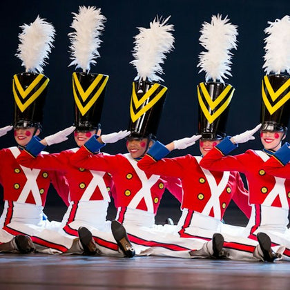 Five performers dressed as toy soldiers in red, white, and black costumes with large hats, sitting in a synchronized pose on stage.