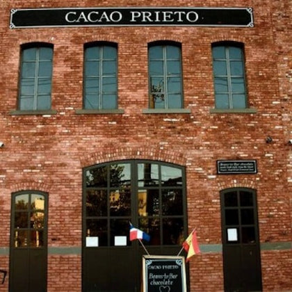 A brick building with the sign 'Cacao Prieto' and a chalkboard outside featuring flags and text about bean-to-bar chocolate.