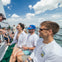 People sitting on a boat, smiling and enjoying drinks under a partly cloudy sky. Water and horizon in the background.