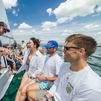 People sitting on a boat, smiling and enjoying drinks under a partly cloudy sky. Water and horizon in the background.