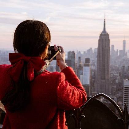 A person with a red bow in their hair and a red coat takes a photo of the New York City skyline, including the Empire State Building.