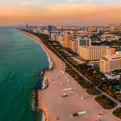 Aerial view of a city coastline with tall buildings, sandy beaches, and turquoise ocean under a sunset sky.