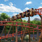 People with raised arms on a red rollercoaster with a Superman logo, under a bright, partly cloudy sky.