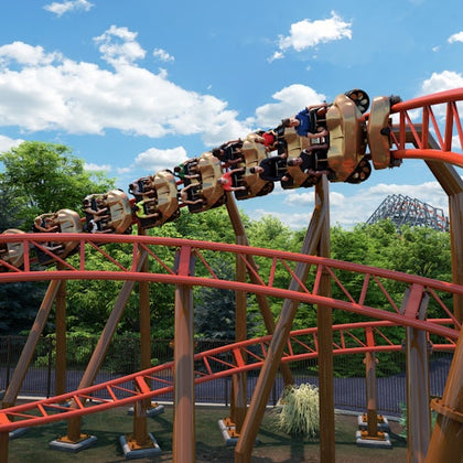 People with raised arms on a red rollercoaster with a Superman logo, under a bright, partly cloudy sky.