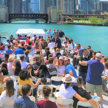 A group of people seated on a boat enjoying a sunny day, with a city skyline and a bridge over a river in the background.
